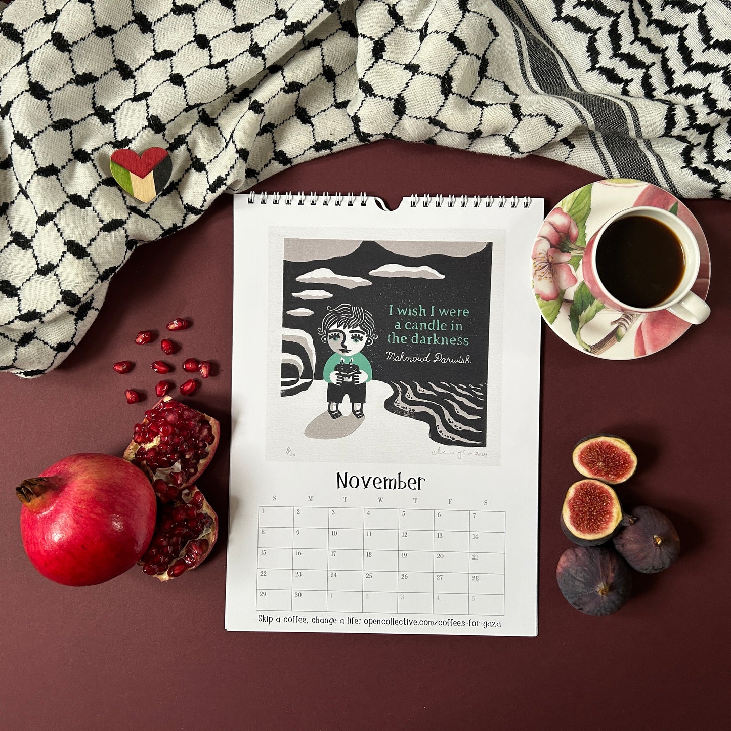 Calendar for November featuring a child holding a candle and quote by Mahmoud Darwish, placed on a surface with fruits and a cup of coffee.